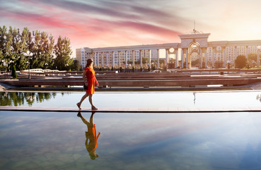 A woman in a red dress walking by a reflective water feature at Republic Square in Almaty, Kazakhstan, with stunning architecture and a vibrant sunset sky in the background.