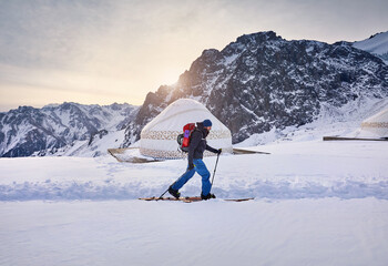 A skier trekking through a snowy landscape, passing traditional yurts with majestic mountains in the background under a serene winter sky.