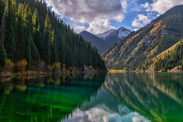 anoramic view of Almaty, Kazakhstan, featuring the Big Almaty lake surrounded by lush green hills and the majestic Tian Shan Mountains under a colorful sunset sky.