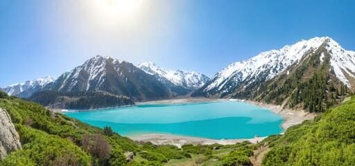 A breathtaking view of the Zailiyskiy Alatau Mountains in Almaty, featuring the turquoise Big Almaty Lake surrounded by lush green vegetation and snow-capped peaks under a bright sunny sky. Keywords: Zailiyskiy Alatau Mountains, Almaty, Big Almaty Lake.