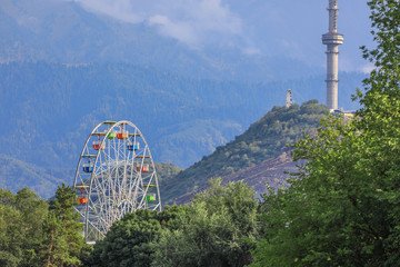 View of Kok-Tobe Hill in Almaty featuring the iconic Ferris wheel surrounded by lush greenery and the Kok-Tobe Tower in the background against a backdrop of majestic mountains