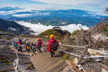 An adventurous group descends a scenic mountain trail in Malaysia, surrounded by breathtaking landscapes of clouds, lush greenery, and rugged terrain. Keywords: Travel Deals: For Couples and Families in Malaysia.