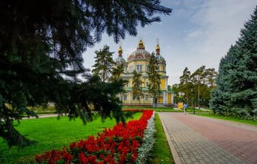 View of Panfilov Park in Almaty showcasing the iconic Zenkov Cathedral surrounded by lush greenery, vibrant red flower beds, and a tree-lined pathway, capturing the park's serene beauty and cultural significance.