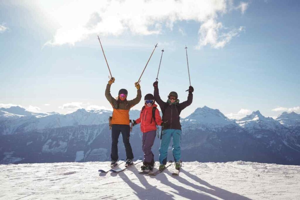 Three skiers in colorful gear celebrating on a snowy mountain peak with ski poles raised, surrounded by stunning alpine scenery.