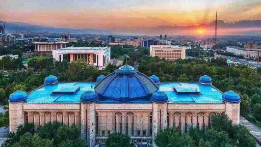 A stunning aerial view of the Central State Museum of Kazakhstan in Almaty, featuring its distinctive blue domes, surrounded by greenery, with a backdrop of the city skyline and a beautiful sunset. image source Divento