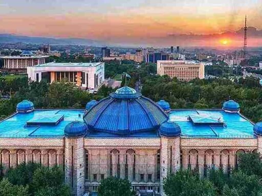 A stunning aerial view of the Central State Museum of Kazakhstan in Almaty, featuring its distinctive blue domes, surrounded by greenery, with a backdrop of the city skyline and a beautiful sunset. image source Divento