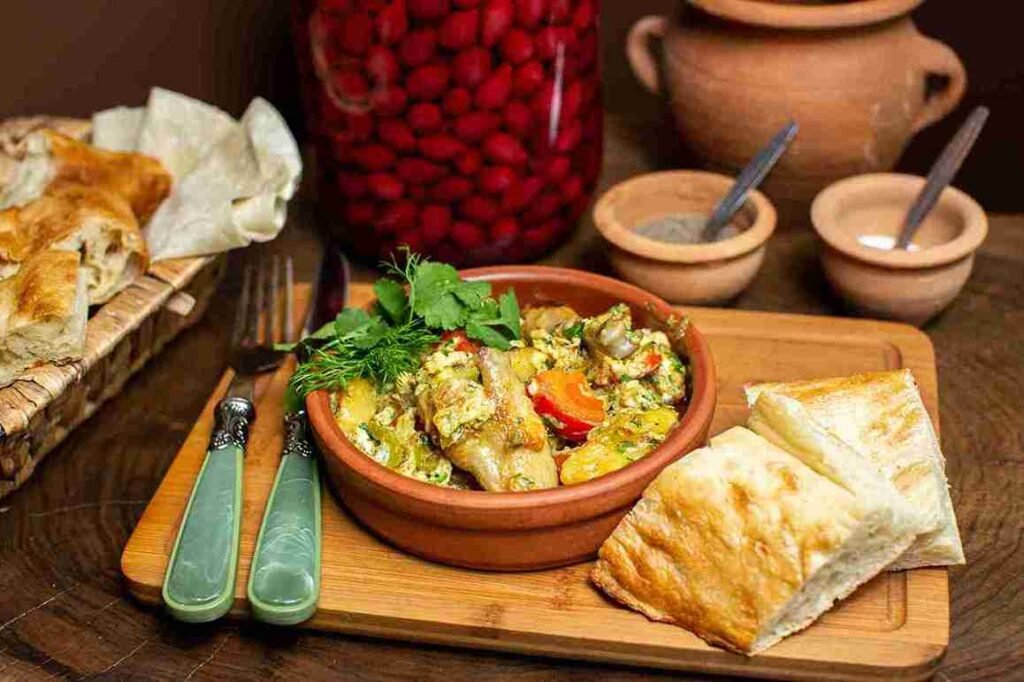 A traditional Almaty dining experience featuring a cooked meal in a brown clay pot, accompanied by fresh bread slices, with herbs and vegetables adding vibrant colors. Tasting local cuisine is one of the best things to do in Almaty for an authentic cultural experience.