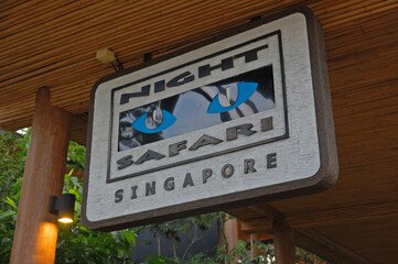 Night Safari Singapore signboard with illuminated design, featuring glowing eyes, set against a wooden background and surrounded by lush greenery. Perfect representation of the world's first nocturnal wildlife park experience.