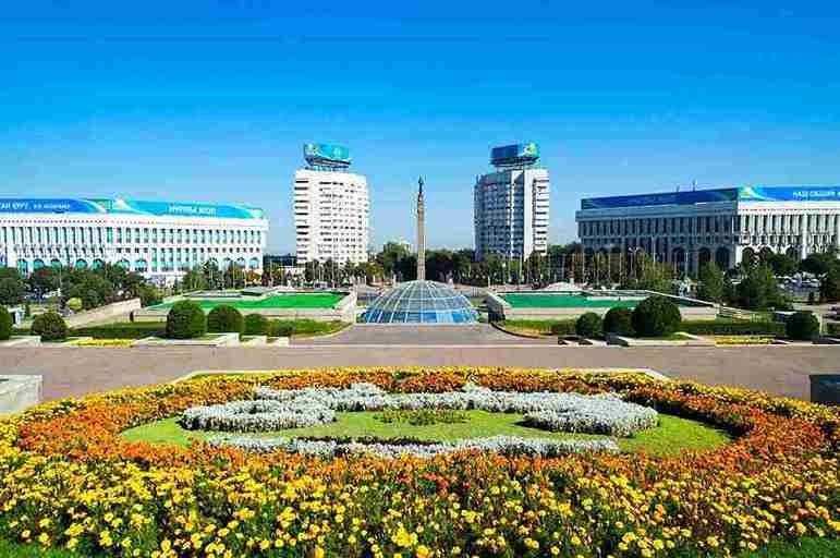 Republic Square in Almaty, Kazakhstan, showcasing vibrant flowerbeds in the foreground, a central glass dome, and iconic administrative buildings with blue rooftops against a clear blue sky