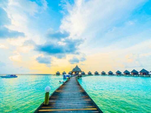 Wooden walkway leading to overwater villas in the Maldives at sunset with turquoise waters and a boat in the distance under a vibrant sky