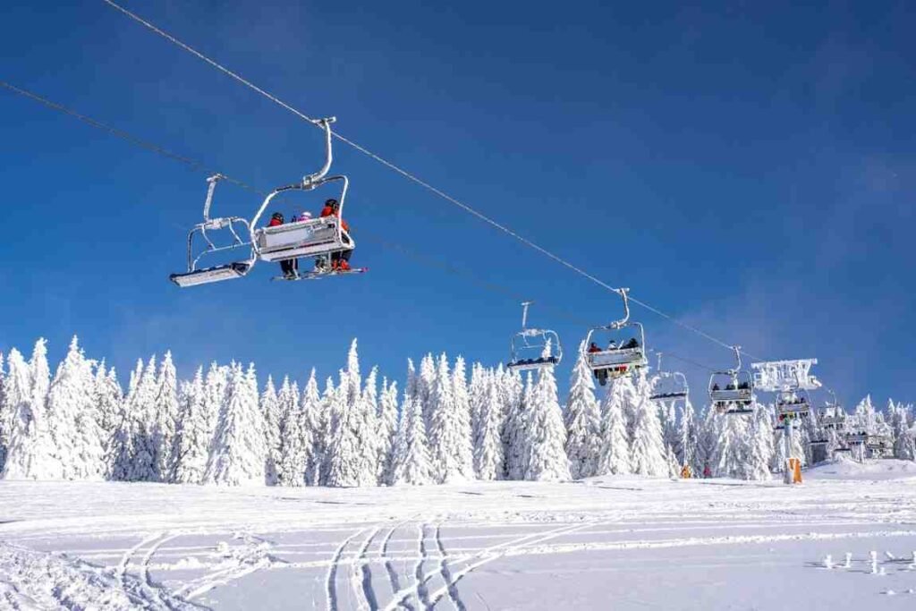 Visitors riding a Shymbulak ski Resort lift at a snowy mountain resort surrounded by frost-covered trees under a clear blue sky.
