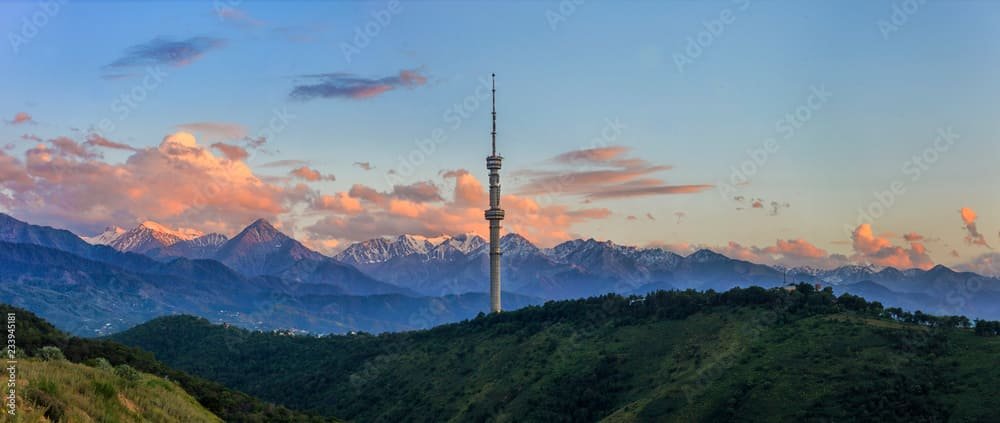 Panoramic view of Almaty, Kazakhstan, featuring the iconic Kok Tobe TV Tower surrounded by lush green hills and the majestic Tian Shan Mountains under a colorful sunset sky.