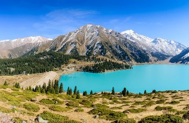 A breathtaking view of Big Almaty Lake in Kazakhstan, surrounded by lush greenery, rolling hills, and snow-capped mountains under a clear blue sky. A serene destination for nature lovers and adventurers. Keywords: Travel to Almaty Kazakhstan.