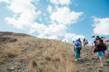 Group of hikers trekking uphill on a dry grassy trail under a bright blue sky with scattered clouds, showcasing an adventurous outdoor activity in a natural landscape.