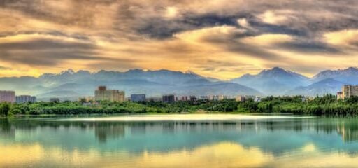 Panoramic view of Alma-Ata City with a serene lake in the foreground, lush greenery, modern architecture, and the majestic Tian Shan Mountains under a vibrant sky.