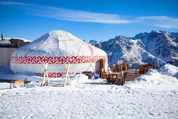 Things to do in Almaty: A traditional yurt set against snow-covered mountains under a clear blue sky, with outdoor seating nearby, showcasing winter activities and cultural experiences in this picturesque destination.