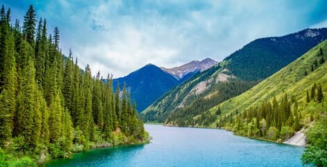 Scenic view of a tranquil lake surrounded by lush green forests and towering mountains in Almaty, Kazakhstan. Reasons to Visit Almaty include its breathtaking natural beauty, serene landscapes, and outdoor adventure opportunities.