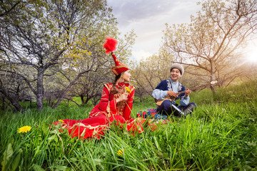 A couple in traditional Kazakh attire enjoying music together in a lush green orchard with blossoming trees and sunlight. Romantic Almaty.