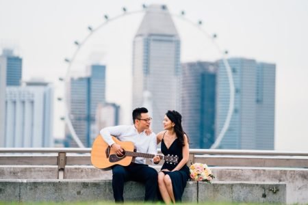 A couple sitting on a concrete bench with a romantic backdrop of the Singapore skyline and the Singapore Flyer. The man, wearing glasses and a white shirt, plays the guitar while looking at the woman in a black dress, who smiles at him. A bouquet of flowers rests beside them, adding to the romantic atmosphere.