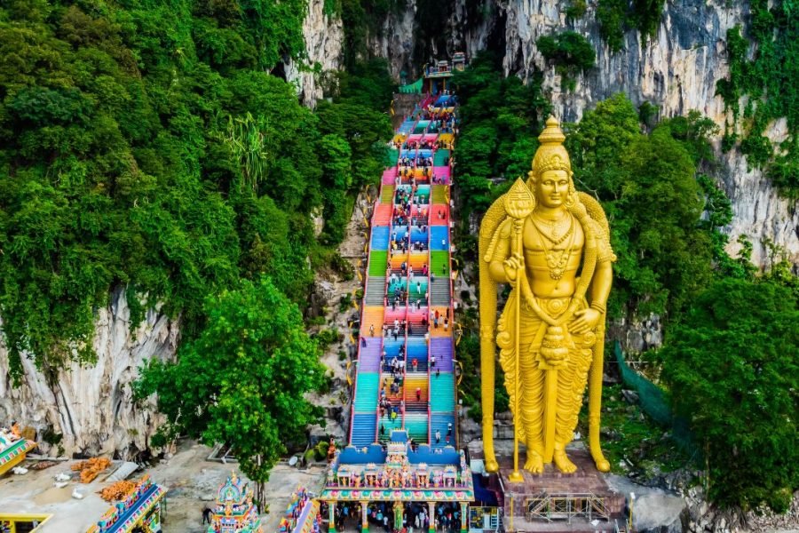 A stunning view of Batu Caves in Malaysia, showcasing the magnificent golden statue of Lord Murugan standing tall beside the vibrant, colorful staircase leading to the ancient limestone caves, surrounded by lush greenery