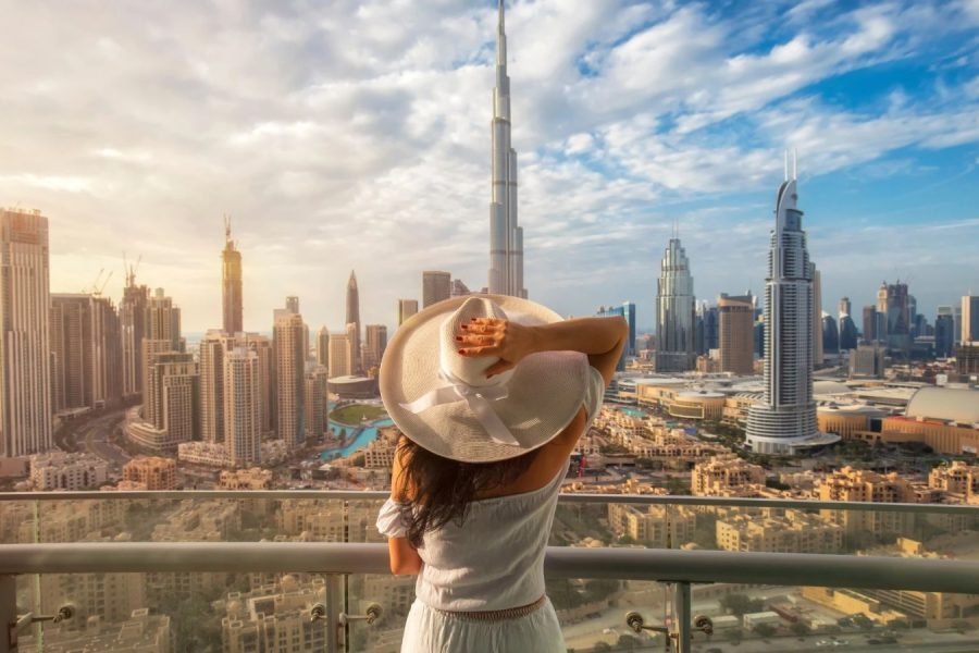 A woman in a white dress and wide-brimmed hat gazes out from a balcony at the stunning Dubai skyline, with the Burj Khalifa prominently rising against a partly cloudy sky.