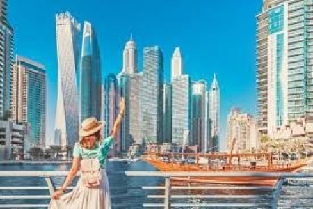 A cheerful woman wearing a hat and backpack waves towards the stunning Dubai Marina skyline. The scene showcases modern skyscrapers, including the iconic Cayan Tower, with a traditional wooden dhow cruising along the canal under a clear blue sky.